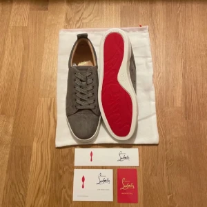 Grå sneakers från Christian Louboutin - Snygga grå sneakers från Christian Louboutin med den ikoniska röda sulan. Skorna har snörning och är tillverkade i mocka, vilket ger en lyxig känsla. Perfekta för den stilmedvetna som vill ha något extra i garderoben.