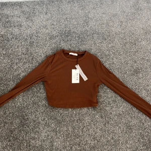 Brun ribbad croppad topp från NA-KD - Säljer en snygg brun ribbad topp från NA-KD. Den är croppad med långa ärmar och har en stilren design. Perfekt för att matcha med höga jeans eller kjol. 🧡