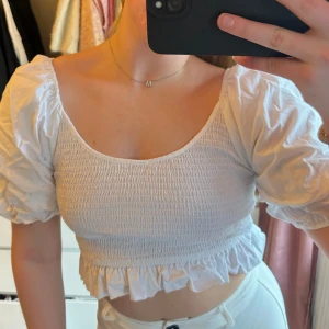 Vit topp från NA-KD - Säljer en söt vit topp från NA-KD med puffiga korta ärmar och smockdetaljer. Toppen är croppad med volangkant nedtill.
