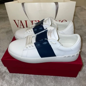 Valentino open  - Snygga vita Valentino sneakers med en bred blå rem över snörningen. Skorna har en stilren design med subtila nitar på hälen och är perfekta för en trendig look.