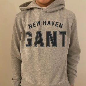 Grå hoodie från GANT - Säljer en stilren grå hoodie från GANT med texten 'NEW HAVEN GANT' i mörkblått på framsidan. Tröjan har en bekväm passform med långa ärmar och en praktisk huva. Perfekt för en avslappnad look!