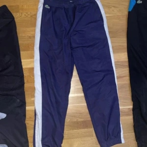 Blå träningsbyxor från Lacoste - Snygga blå träningsbyxor från Lacoste med vita sidopaneler och elastisk midja med snörning. Perfekta för sportiga aktiviteter eller en avslappnad stil. Den ikoniska Lacoste-loggan pryder framsidan.