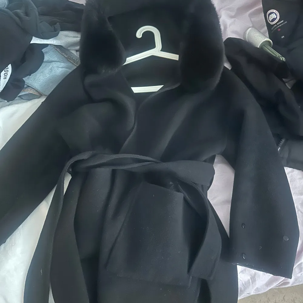 Stilren och elegant svart kappa med lyxiga pälsdetaljer på huvan och ärmarna. Ärmpälsen är avtagbar, vilket gör det enkelt att anpassa kappan efter tillfälle och stil. Knappt använd. Xs/S. Takit.