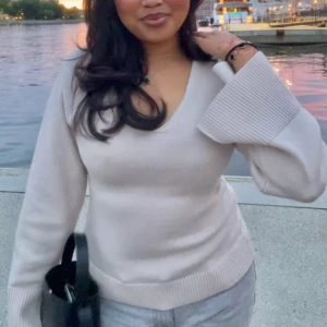 Beige stickad tröja med v-ringning - Säljer en stilren beige stickad tröja med v-ringning. Tröjan har långa ärmar och ribbade detaljer vid ärmslut och nederkant. Perfekt för en avslappnad och trendig look.