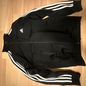 Svarta träningsbyxor från Adidas - Snygga svarta träningsbyxor från Adidas med de klassiska tre vita ränderna längs sidorna. Byxorna har en elastisk midja och en liten Adidas-logga på ena benet. Perfekta för träning eller en avslappnad stil.