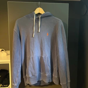 Blå hoodie från Polo Ralph Lauren - Säljer en blå hoodie från Polo Ralph Lauren i storlek M. Tröjan har vit snörning i huvan, klassisk känguruficka och den ikoniska orangea Polo-loggan broderad på bröstet. Perfekt för dig som gillar stilren och avslappnad look. Sliten i sömmar.