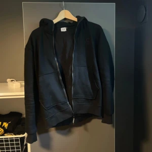 Svart CP Company hoodie med goggles - Svart hoodie från CP Company med ikoniska goggles i huvan. Tröjan har dragkedja framtill, två stora fickor och en relaxed passform. Perfekt för dig som vill sticka ut med streetwear-vibe. Materialet är mjuk bomull och huvan är riktigt fet. Tyvärr är zippen trasig lite nedanför halsen.