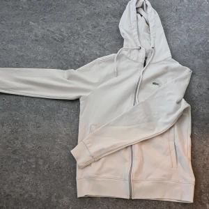 Vita mjukisbyxor från Lacoste - Snygga vita mjukisbyxor och kåfta från Lacoste med klassisk krokodillogga på benet. Byxorna har resår och snörning i midjan samt muddar vid bensluten för en skön passform. Perfekta för en chill och sportig look.