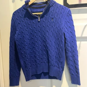 Blå stickad tröja Polo Ralph Lauren - Kabelstickad blå tröja från Polo Ralph Lauren med half zip och hög krage. Tröjan har ribbade muddar och ärmar, samt diskret broderad logga på bröstet. Perfekt för dig som gillar klassisk och sportig stil.