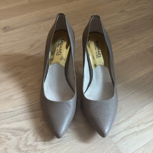 Beige pumps från Michael Kors - Snygga pumps från Michael Kors i beige skinn med spetsig tå och låg klack. Skorna har en elegant struktur och guldfärgade detaljer baktill. Perfekta för dig som vill ha en stilren och klassisk look.