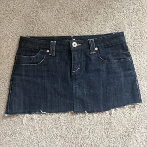 Mörk jeanskjol med fransig kant - Snygg mörkblå jeanskjol från YMI med rå, fransig nederkant och klassiska fickor både fram och bak. Kjolen har bälteshällor och kontrastsömmar som ger en cool vibe. Perfekt för dig som gillar en edgy och avslappnad stil.