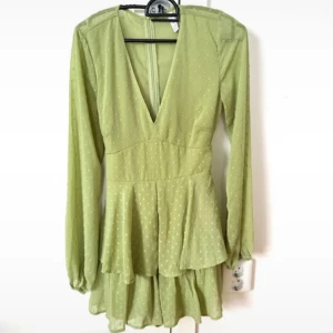 Ljusgrön kortklänning med volanger - Superfin ljusgrön kortklänning med v-ringning, långa ärmar och volangdetaljer. Klänningen har små prickar i tyget, knytband i midjan och är tillverkad i ett lätt, luftigt material. Perfekt för vår och sommar!