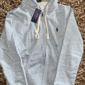 Grå hoodie från Polo Ralph Lauren - Snygg ljusgrå hoodie från Polo Ralph Lauren med dragkedja, huva och vita snören. Broderad marinblå logga på bröstet och känguruficka framtill. Perfekt för chill dagar och enkel att matcha med jeans eller joggers.