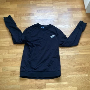 Svart långärmad tröja EA7 Armani - Svart långärmad tröja från EA7 Emporio Armani med vit logga på bröstet och diskreta ränder i nacken. Klassisk rund hals och mjukt material som känns skönt mot huden. Perfekt för dig som gillar stilren och sportig design.