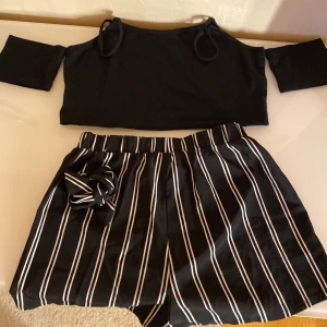 Set: svart top och randiga shorts med bälte - Snygg svart top och svarta shorts med vita vertikala ränder och elastisk midja. Shortsen har ett bälte, perfekta för varma dagar. Materialet känns lätt och luftigt, vilket gör dem grymt sköna på sommaren.