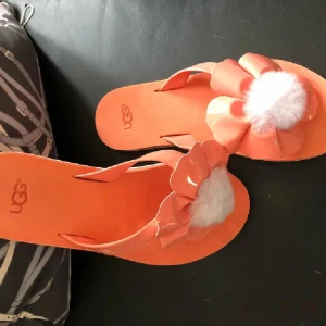UGG sandaler med blomdetalj orange - Snygga orange sandaler från UGG med stor blommig detalj och fluffig vit mitt på ovansidan. Sulan är ljuslila med mönster och remmarna är i matchande orange syntetmaterial. Perfekta för sommardagar när du vill sticka ut lite extra.