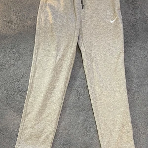 Grå mjukisbyxor från Nike - Snygga grå mjukisbyxor från Nike med dragsko i midjan och två sidofickor. Byxorna har en klassisk, avslappnad passform . Perfekta för chill eller träning, med diskret Nike-logga broderad på benet. Storlek L men käns som M. Längden är 102 cm
