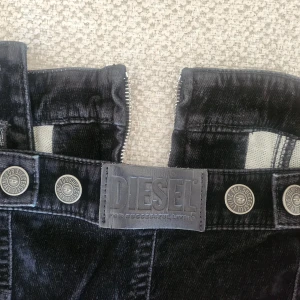 Svart manchesterklänning från Diesel - Supersnygg svart kortklänning från Diesel i mjuk manchester med axelband och markerad midja. Klänningen har dragkedja framtill, coola sömdetaljer och fickor både fram och bak. Man kan dessutom ta av axelbanden. Perfekt för dig som vill sticka ut med en edgy look. Orginalpriset var runt 2000kr