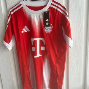 Bayern München Musiala matchtröja - Röd och vit Bayern München fotbollströja från Adidas med Musiala och nummer 10 på ryggen. Klassisk design med vita detaljer, klubbmärke och sponsorlogga framtill. Korta ärmar och tillverkad i lätt funktionsmaterial, perfekt för match eller träning.