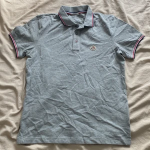 Grå pikétröja från Moncler - Snygg grå pikétröja från Moncler med klassisk krage och knäppning framtill. Tröjan har Moncler-logga på bröstet och detaljer i rött, vitt och blått på krage och ärmslut. Perfekt för en clean och stilren look.