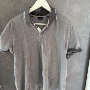 Grå pikétröja från Hugo Boss - Snygg grå pikétröja från Hugo Boss med diskret logga broderad på bröstet. Tröjan har klassisk krage i mörkare grå nyans och vita detaljer längs krage och ärmslut. Perfekt för en clean och stilren look. (Liten i storleken)