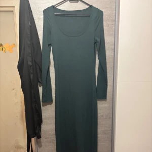 Mörkgrön långärmad långklänning - En stilren mörkgrön långklänning med lång ärm och rundad urringning. Klänningen har en slim passform och är tillverkad i ett mjukt, stretchigt material som sitter snyggt på kroppen. Perfekt för dig som gillar minimalistisk och clean stil.