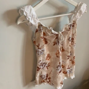 Blommig spetslinnetopp med volang - Romantisk linnetopp med blommigt mönster i beige, brunt och vitt. Toppen har breda axelband i vit spets, volangkant och söt knytning framtill. Materialet är lätt och luftigt, perfekt för varma dagar. Snyggt broderad spetsdetalj längs kanterna för extra vintagekänsla.