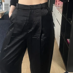 Svarta kostymbyxor i sidenblandning, H&M Premium - Snygga svarta kostymbyxor från H&Ms Premiumkollektion i sidenblandning. Byxorna har hög midja, raka ben och en glansig finish som ger en lyxig känsla. Klassisk passform med hällor för bälte och fickor i sidorna. Tyvärr aldrig använda pga sensory issues. Ganska små i storleken, beroende på midjemått är dom nog 36/38, jag är normalt en 36a. 