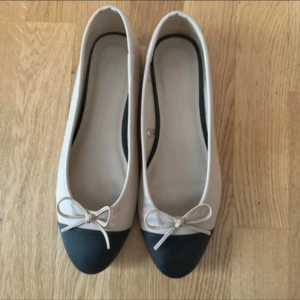 Beige ballerinaskor med rosett H&M - Snygga ballerinaskor från H&M i beige med svart tå och söt rosettdetalj framtill. Skorna har platt sula och rund tå, tillverkade i syntetmaterial. Perfekta för en stilren och klassisk look.