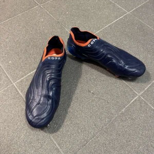Adidas Copa blå fotbollsskor - Mörkblå Adidas Copa fotbollsskor med orange detaljer och texten FUSIONSKIN på sidan. Skorna har en strumpliknande öppning och mönstrad ovandel i skinn. Sulan är blå med fasta dobbar för gräsplan. Snygg och modern design för dig som vill sticka ut på planen. skorna är använda några gånger bara