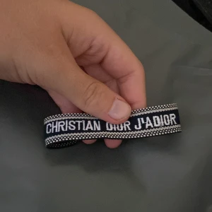 Christian Dior J'Adior armband - Snyggt vävt armband från Christian Dior med texten 'J'Adior' i vitt på mörkblå botten. Armbandet har justerbar knytning med tofsar i matchande färger. Perfekt accessoar för att lyfta din outfit med en lyxig touch.