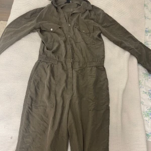 Olivgrön jumpsuit från H&M - Snygg olivgrön jumpsuit från H&M i storlek 36. Modellen har långa ärmar, krage, knappar framtill och två bröstfickor med lock. Skriv för fler frågor om mått eller annat💗