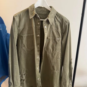 Olivgrön jeans jacka - Snygg olivgrön jeansjacka med slitna och lagade detaljer för en cool, avslappnad look. Skjortan har två bröstfickor, knappar framtill och klassisk krage. Perfekt att bära öppen över en t-shirt eller stängd för en mer stilren vibe.