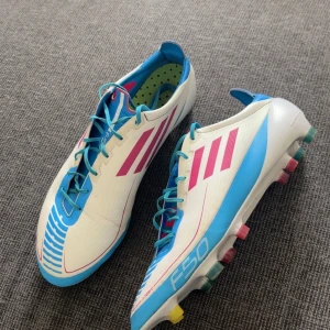 Adidas F50 Adizero fotbollsskor blå/vit - Säljer ett par Adidas F50 Adizero fotbollsskor i vitt med blå och rosa detaljer. Skorna har snörning, färgglada dobbar och en lätt, sportig design. Perfekta för dig som vill sticka ut på planen med stilrena och snabba skor.