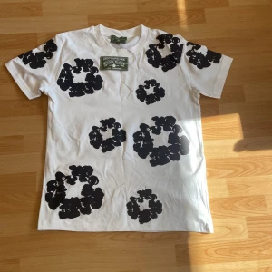 Vit t-shirt med svarta blommor Denim Tears - Säljer en vit t-shirt från Denim Tears med stora svarta blommiga tryck över hela plagget. Klassisk passform med rund hals och korta ärmar. Materialet är mjuk bomull som känns skönt mot huden. Perfekt statement-plagg för dig som gillar unik streetwear.