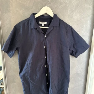 Mörkblå kortärmad skjorta SMDG - Stilren mörkblå skjorta från SMDG i luftig bomull. Skjortan har korta ärmar, klassisk krage och vita knappar framtill. Perfekt för varma dagar när du vill vara både bekväm och snygg.