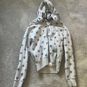 Grå hoodie med stjärnor från D&G - Snygg grå hoodie från D&G med svarta stjärnor över hela tröjan och stor D&G-logga framtill. Tröjan har luva, croppad passform och långa ärmar. Har aldrig använt men dock köpt second hand