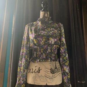 Blommig blus från Vero Moda  - Supersöt blommig blus från Vero Moda i storlek XS. Blusen har hög krage, långa ärmar och smockad midja som ger en snygg siluett. Färgerna är lila, gul, vit och grön med blommigt mönster. Perfekt för dig som gillar färg och detaljer! Knappt använd och inga defekter vad jag kan se. 