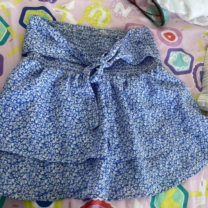 Blåvit blommig kortkjol med knyt - Supersöt blå och vit kortkjol med blommigt mönster och smock i midjan. Kjolen har en volangig känsla och ett knytband framtill som ger extra detalj. Perfekt för varma dagar och lätt att matcha med en enkel topp.