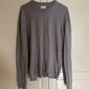 Grå stickad tröja från A days march  - En stilren grå stickad tröja från A days march med rund halsringning och långa ärmar. Tröjan har en klassisk passform och är tillverkad i mjukt material som passar perfekt för lager-på-lager under kyliga dagar. Storlek M 