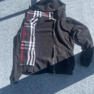 Svart Burberry hoodie - Svart hoodie från Burberry i storlek M med dragkedja och klassiskt rutig mönster i svart, vitt och rött på insidan. Jackan har huva, ribbade muddar och känguruficka framtill. Perfekt för dig som vill ha en stilren streetlook med lyxig touch. Kom privat vid fler funderingar 