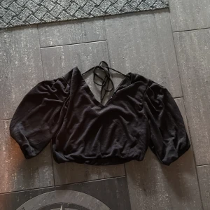 Svart croppad blus med puffärm - Svart blus med croppad passform, v-ringning och knytband i nacken. Toppen har puffiga trekvartsärmar som ger en trendig siluett. Perfekt för dig som gillar stilrena och coola plagg med det lilla extra.
