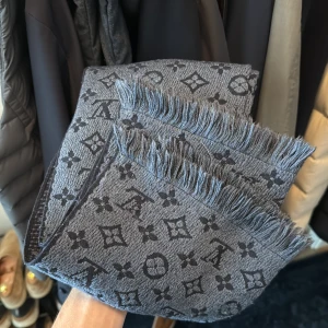 Grå Louis Vuitton halsduk i ull - Snygg grå halsduk från Louis Vuitton med ikoniskt monogrammönster i mörkare grå toner. Halsduken är i 100% ull och har fransade kanter för en extra cool vibe. Perfekt accessoar för att lyfta din vinterstil.