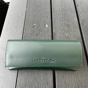 Svarta solglasögon från Lacoste - Stilrena och helt nya svarta solglasögon från Lacoste med rektangulära bågar i matt finish. Snygg metallram och mörka glas, perfekt för en clean och modern look. Kommer med grön Lacoste-fodral. Diskret logga på skalmen för extra premiumkänsla. Fick inte lämna tillbaka de då jag råkade köpa 2, går ordinarie för 2300. Pris är inte ristat i sten