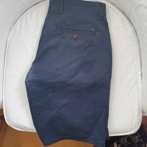 Mörkblå chinos från Tommy Hilfiger  - Mörkblå chinos shorts från Tommy Hilfiger med klassisk passform. Byxorna har sidofickor, bakficka med knapp och diskret Hilfiger-logga ovanför bakfickan samt vid vänster ficka. De är ganska använda som man kan se på bilderna därav det lägre priset! 