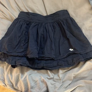 Mörkblå volangkjol - Supersöt mörkblå kortkjol från Abercrombie & Fitch med volang och bred midja. Kjolen har en liten vit logga broderad längst ner och är perfekt för dig som gillar en luftig och lätt stil. Passar till sneakers eller boots för en chill vibe.