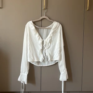Vit blus med volang och knyt - Supersöt vit blus i tunn, lätt transparent bomull med diskreta ränder. Blusen har långa ärmar med volangdetaljer och knytband vid ärmslut. Framtill finns volang längs v-ringningen och ett knytband. Perfekt för en romantisk look.