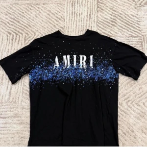 Svart Amiri t-shirt med blått stänkmönster - Snygg svart t-shirt från Amiri med stort vitt logotryck framtill och blått stänkmönster över bröstet och ryggen. Klassisk passform med rund hals och korta ärmar. Perfekt för dig som gillar streetwear och vill sticka ut lite extra.