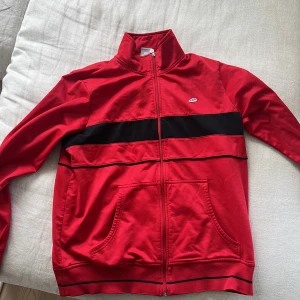 Röd Nike track jacket med svart detalj - Röd track jacket från Nike med svart horisontell rand över bröstet. Jackan har dragkedja hela vägen, ståkrage och två fickor framtill. Perfekt för en sportig och avslappnad stil. Klassisk Nike-logga på bröstet och snygga sömdetaljer.