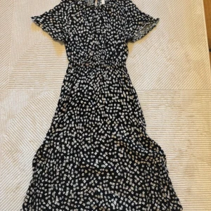 Klänning  - Supersöt svart midiklänning med vita blommor och korta ärmar. Klänningen har en lätt utställd form och knäppning bak i nacken. Perfekt för dig som gillar en feminin och avslappnad stil. Storlek XS men är mer som S